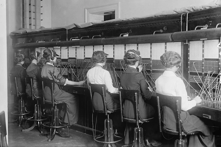 Black-and-white photo of six women wearing headphones and wearing old-fashioned clothing plugging cables into sockets