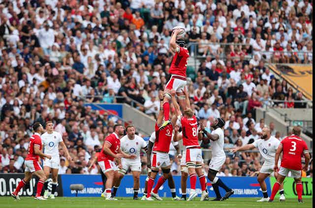 Un groupe de rugbymen en rouge hisse un joueur au-dessus d'un groupe de rugbymen en blanc. 