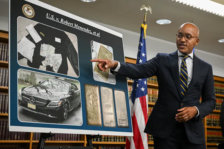 A man in a suit pointing at a poster board with various photos on it.