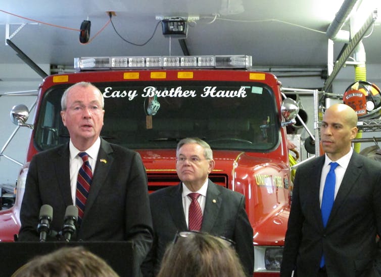 Three men in suits, standing in front of a fire engine.