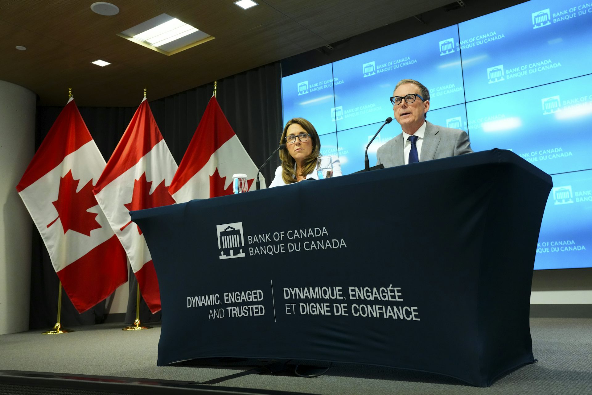 A man and a woman, both wearing glasses, sit on stage behind a Bank of Canada banner. Microphones are in front of them and a row of Canadian flags is behind them.