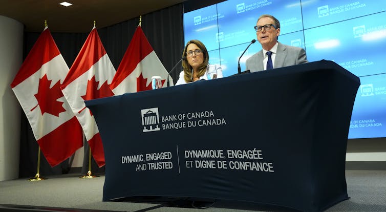 A man and a woman, both wearing glasses, sit on stage behind a Bank of Canada banner. Microphones are in front of them and a row of Canadian flags is behind them.