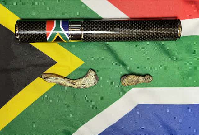 Un tubo negro y plateado, con una pequeña bandera sudafricana en el lateral, y dos huesos recostados sobre una gran bandera sudafricana.