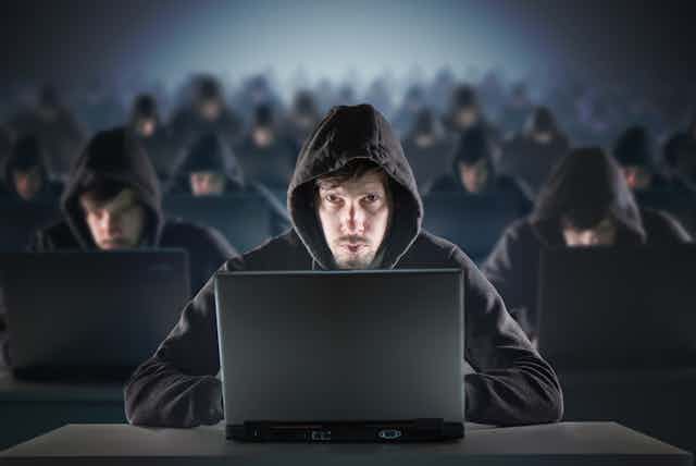 dozens of men wearing black hoodies sitting in front of laptops.