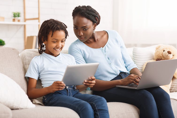 A young girl holding an ipad sits next to her mother on a sofa. The mother has her own laptop but is looking at her daughter's iPad screen.