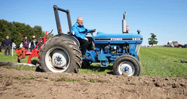 A rotund man with slicked back white hair drives a tractor.