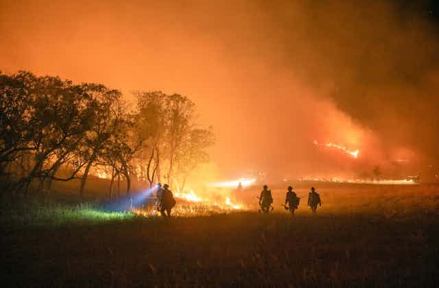 Silhouettes of five firefighters, one with a headlamp lighting the way, walking through a wooded field with fire burning in the background.
