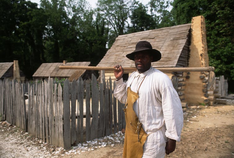 A Black man dressed in an apron and carrying a stick is walking past a small house.