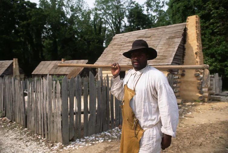 A Black man dressed in an apron and carrying a stick is walking past a small house.