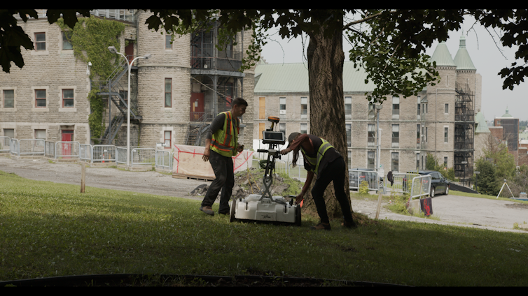Men using ground penetrating radar equipment in a grassy area near a building.