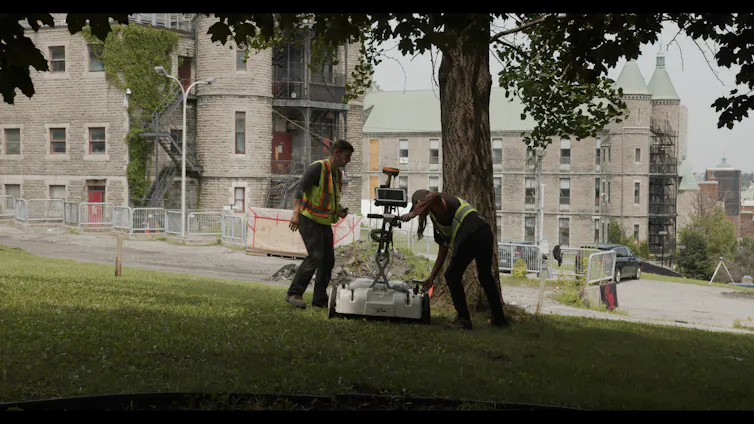 Men using ground penetrating radar equipment in a grassy area near a building.