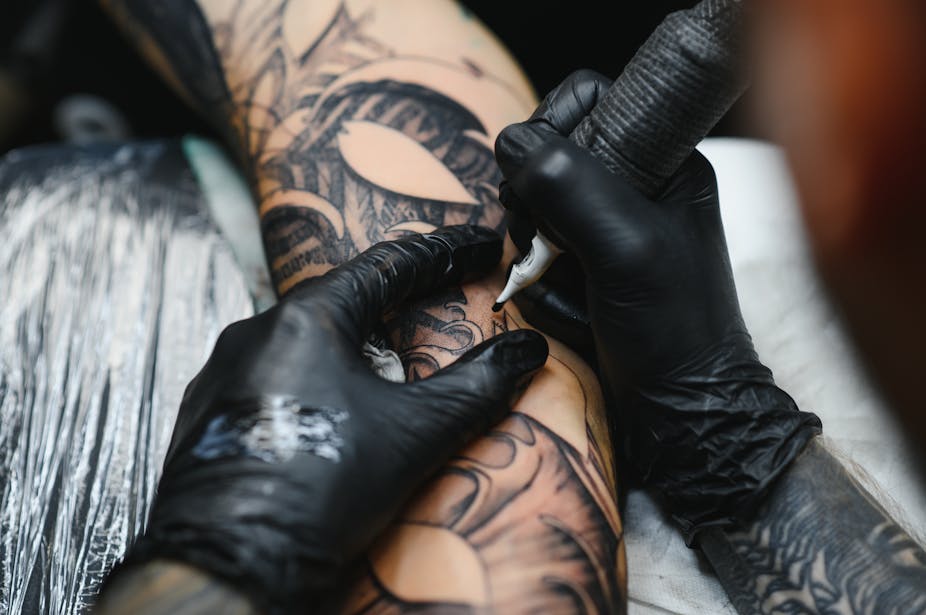 A tattoo artist wearing black gloves uses a needle to tattoo a person's skin with black ink.