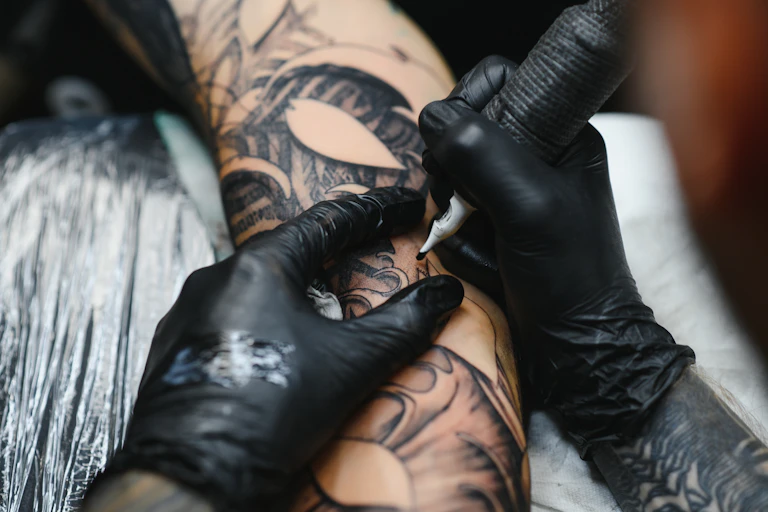 A tattoo artist wearing black gloves uses a needle to tattoo a person's skin with black ink.
