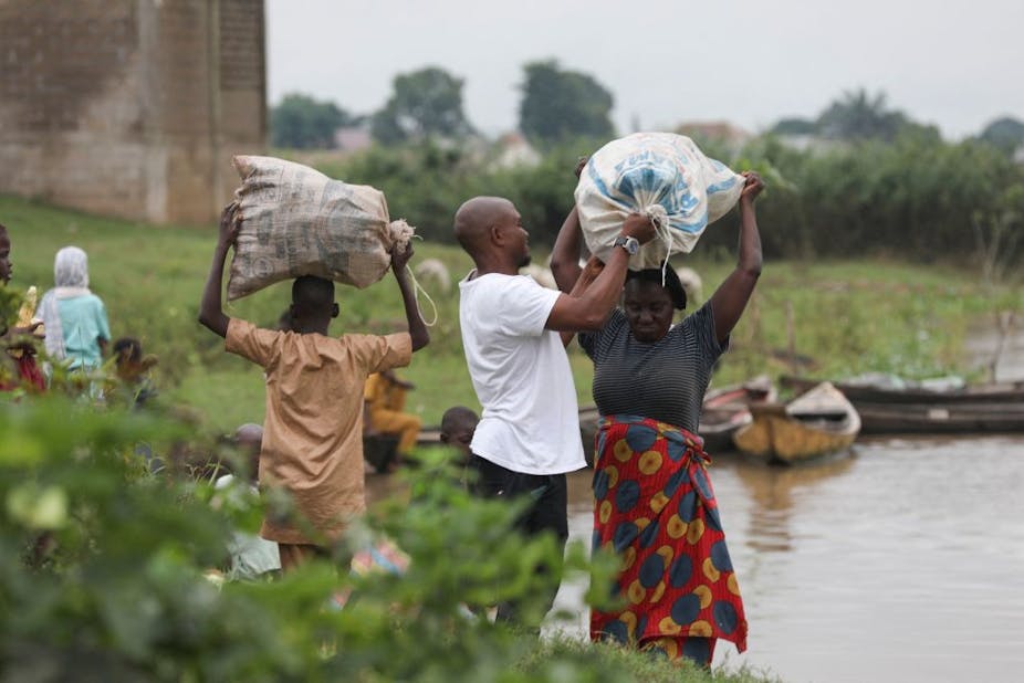 A man helps a woman to balance a large, full plastic bag on her head. They are standing alongside a river and others are seen in the distance, also carrying full bags on their heads