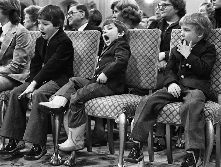 A black and white photo shows three young boys sitting in a row, two yawning.