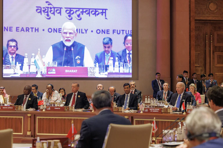 Dignitaries in suits from around the word seated in a conference room while a large screen depicts a bearded man speaking into a microphone.