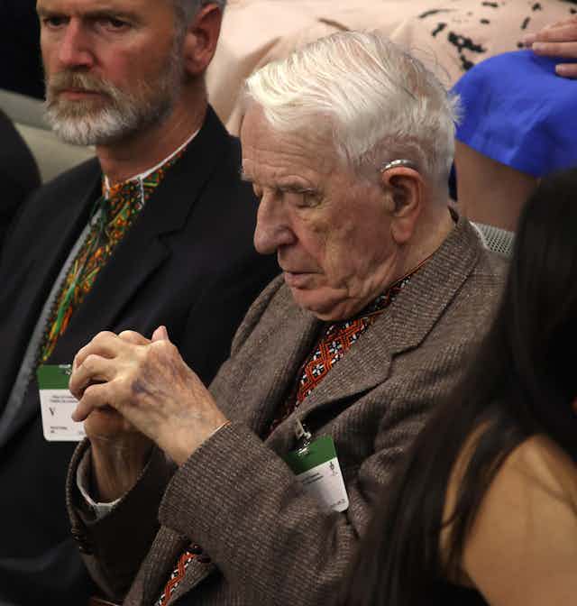 A white-haired man sits with his head bowed and his hands clasped.
