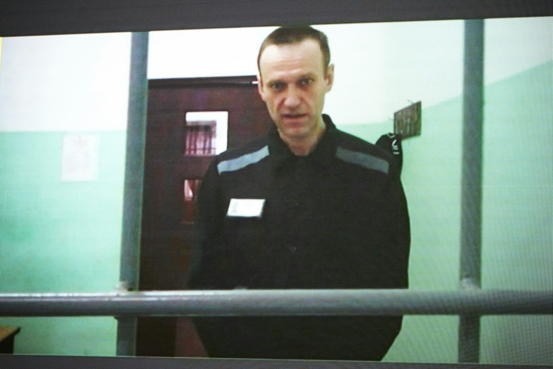 A thin man with dark hair dressed in dark clothing stands in a room behind barricades.