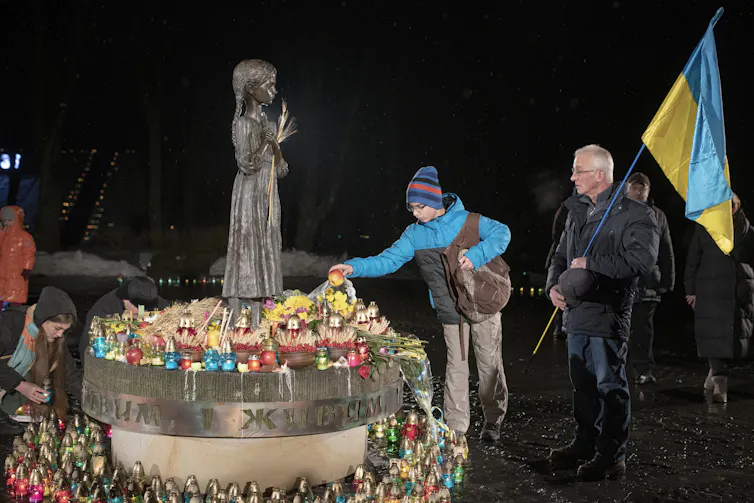 A child in a park lays flowers at a monument. The man behind him is holding a blue and yellow flag.