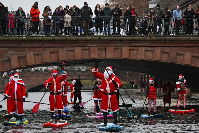 Half a dozen people in red and white fuzzy suits paddleboard beneath a bridge as a crowd watches above.