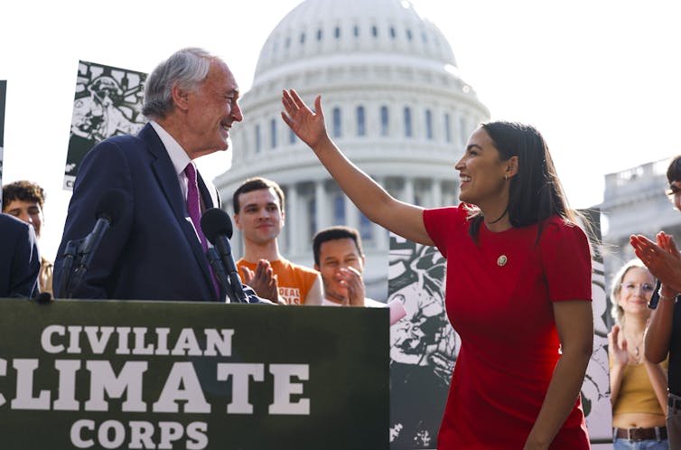 Two politicians celebrate in front of the U.S. Capitol building with a sign saying 'civilian climate corps.'