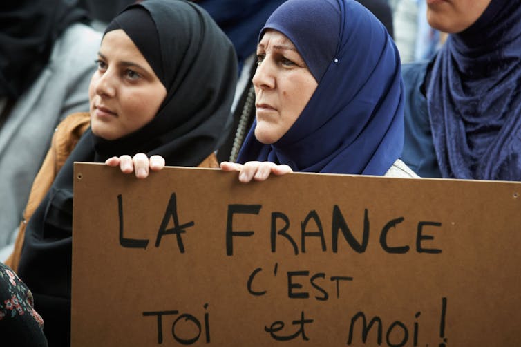 Two women in headscarves hold a cardboard sign written in black marker.