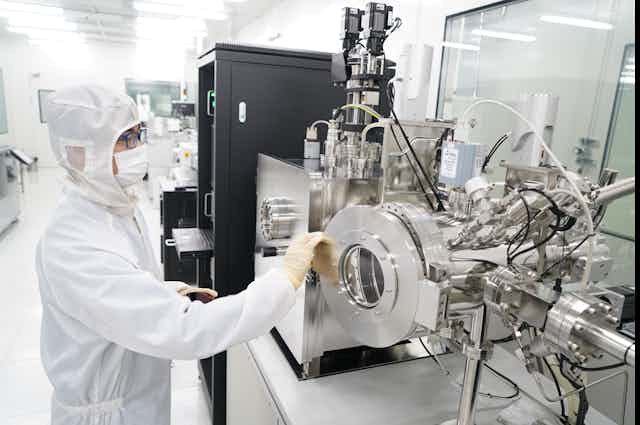 A person in a full body covering wearing glasses, a facemask and rubber gloves opens a small round door on a shiny steel piece of equipment in a laboratory