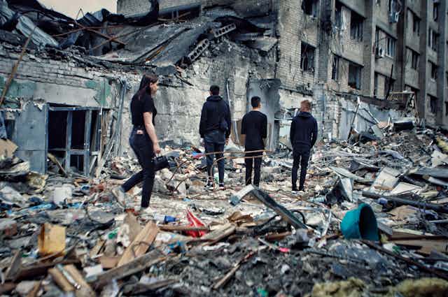Four people stand amid piled debris on a street next to a partially collapsed building.