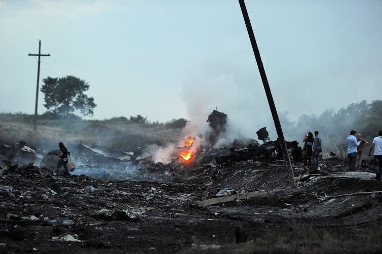 A flame burns in wreckage near a group of people at the site of a destroyed airplane.