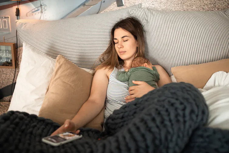 A woman lies on a bed, holding a sleeping newborn.