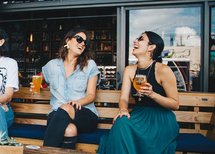 Two women sit on a bench at a bar, drinking and laughing.