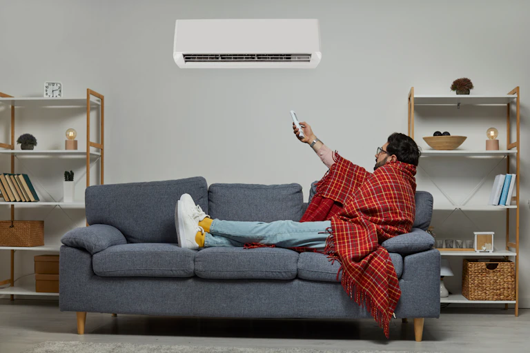 Man wrapped in a blanket on a couch adjusts air conditioner temperature