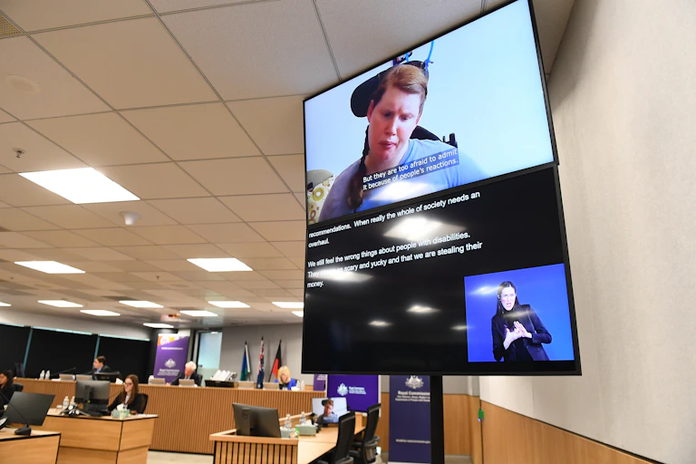 woman in wheelchair speaks on video screen in a hearing room
