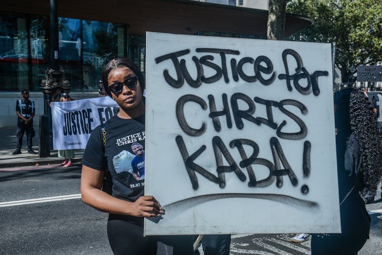 A woman wearing sunglasses holds a sign reading Justice for Chris Kaba! in spraypaint