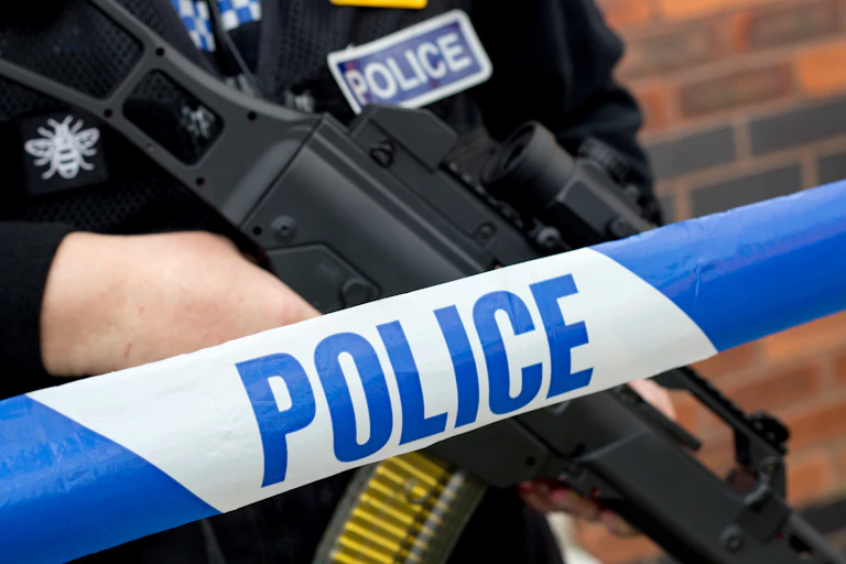 A blue and white police crime scene tape with an armed officer standing behind it