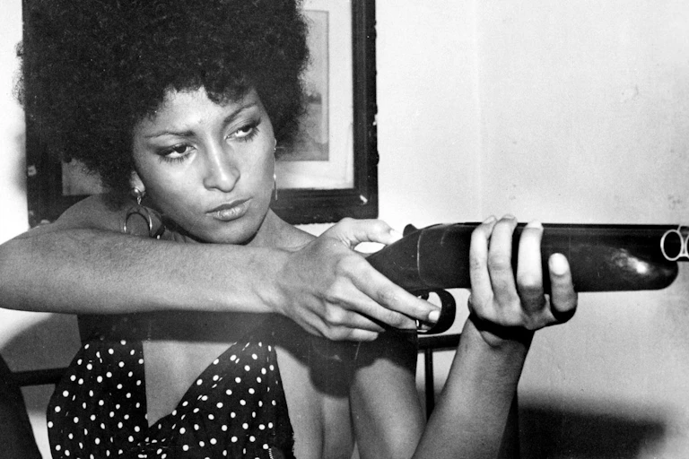 A black woman, eyes narrowed, holding a sawn-off shotgun.