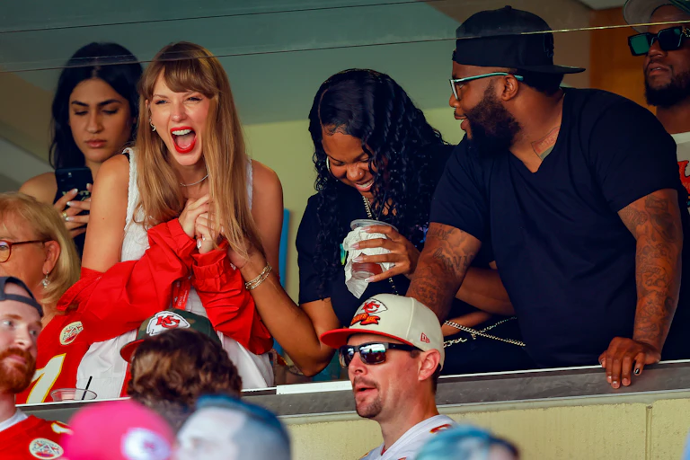 Blonde-haired woman wearing red jacket laughs and claps from a luxury box at a football game.