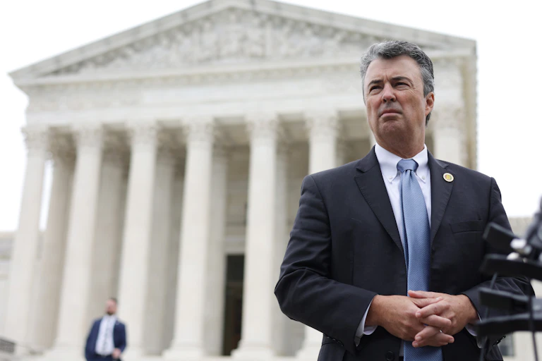 A middle-aged white man dressed in a business suit stands in front of a white building.