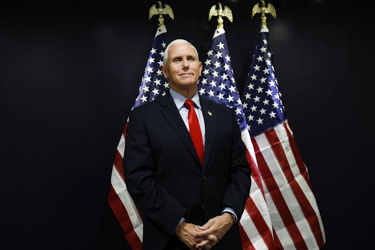 A man dressed in a blue suit, white shirt and red tie posing in front of three American flags.