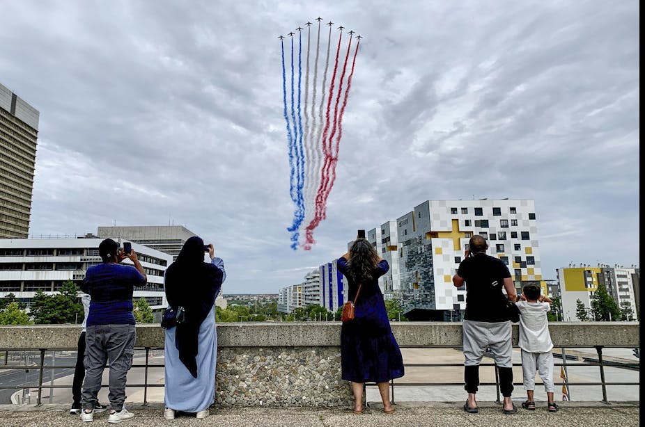 Four adults and a child, including a woman in a headscarf and long dress, take photos as a row of jets flies overhead with plumes of smoke in the colors of the French flag.