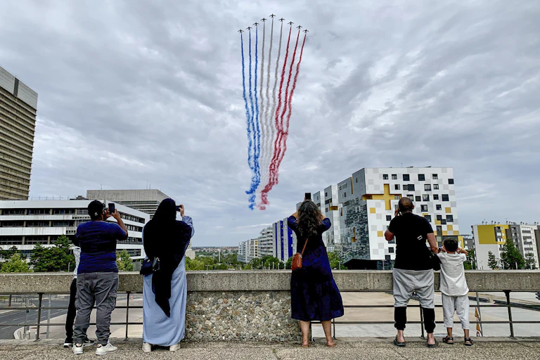 Four adults and a child, including a woman in a headscarf and long dress, take photos as a row of jets flies overhead with plumes of smoke in the colors of the French flag.