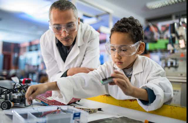 A young boy wearing a lab coat holds a computer part as his teacher, also in a lab coat, stands nearby.
