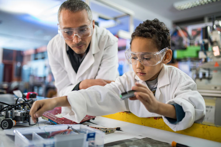 A young boy wearing a lab coat holds a computer part as his teacher, also in a lab coat, stands nearby.