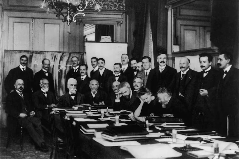 A group of men in suits sit around a table covered in books, with a chandelier above and an easel in the back. The photo is in black and white.