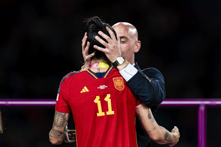 A man in a suit grabs a female soccer player by the head while kissing her.