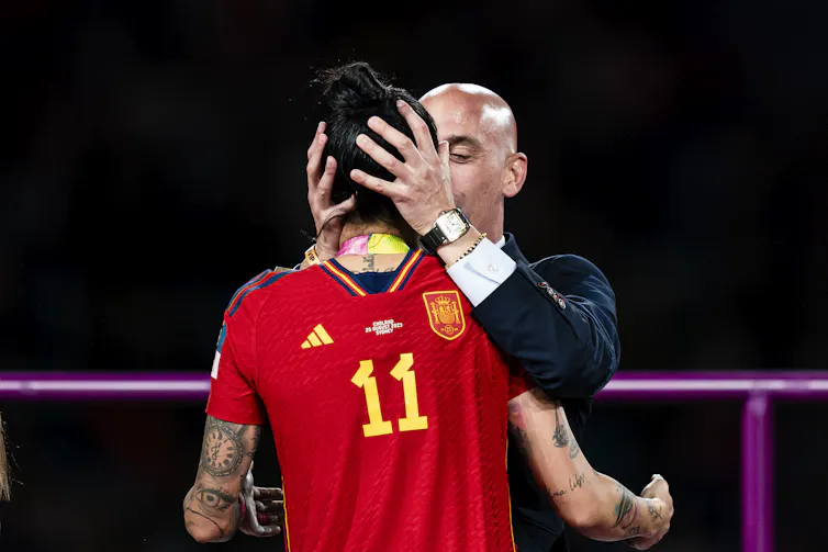 A man in a suit grabs a female soccer player by the head while kissing her.