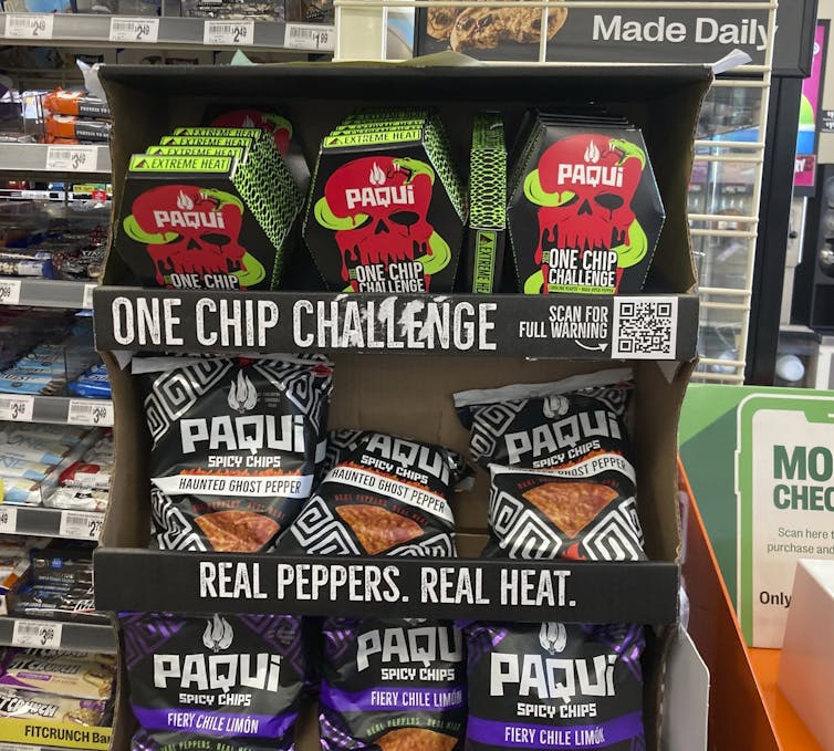 A cardboard display at a gas station reading 'One Chip Challenge Real Peppers Real Heat' with several bags and boxes of 'Paqui' brand chips.