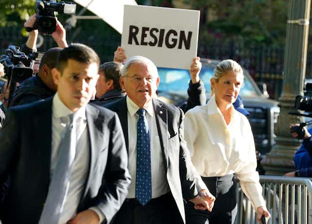 As a gray haired man holds hands with a woman and walks on a crowded sidewalk, someone holds up a white sign with black writing behind his head that reads, “‘Resign.”