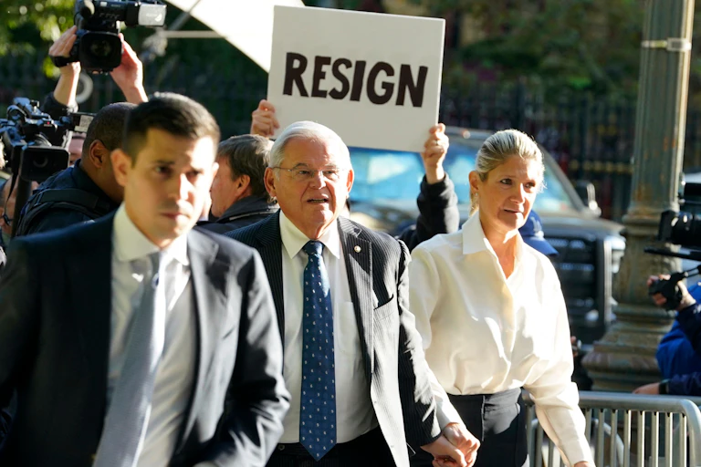As a gray haired man holds hands with a woman and walks on a crowded sidewalk, someone holds up a white sign with black writing behind his head that reads, “‘Resign.”