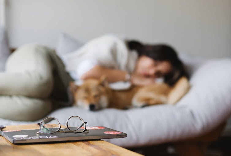 A woman naps with a dog. Spectacles are folded on a book.